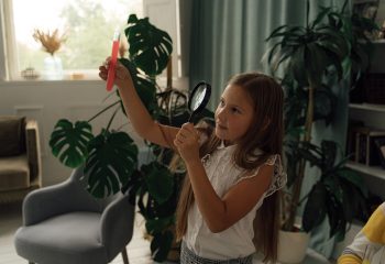 A young girl examining a test tube with a magnifying glass in a cozy room setting.